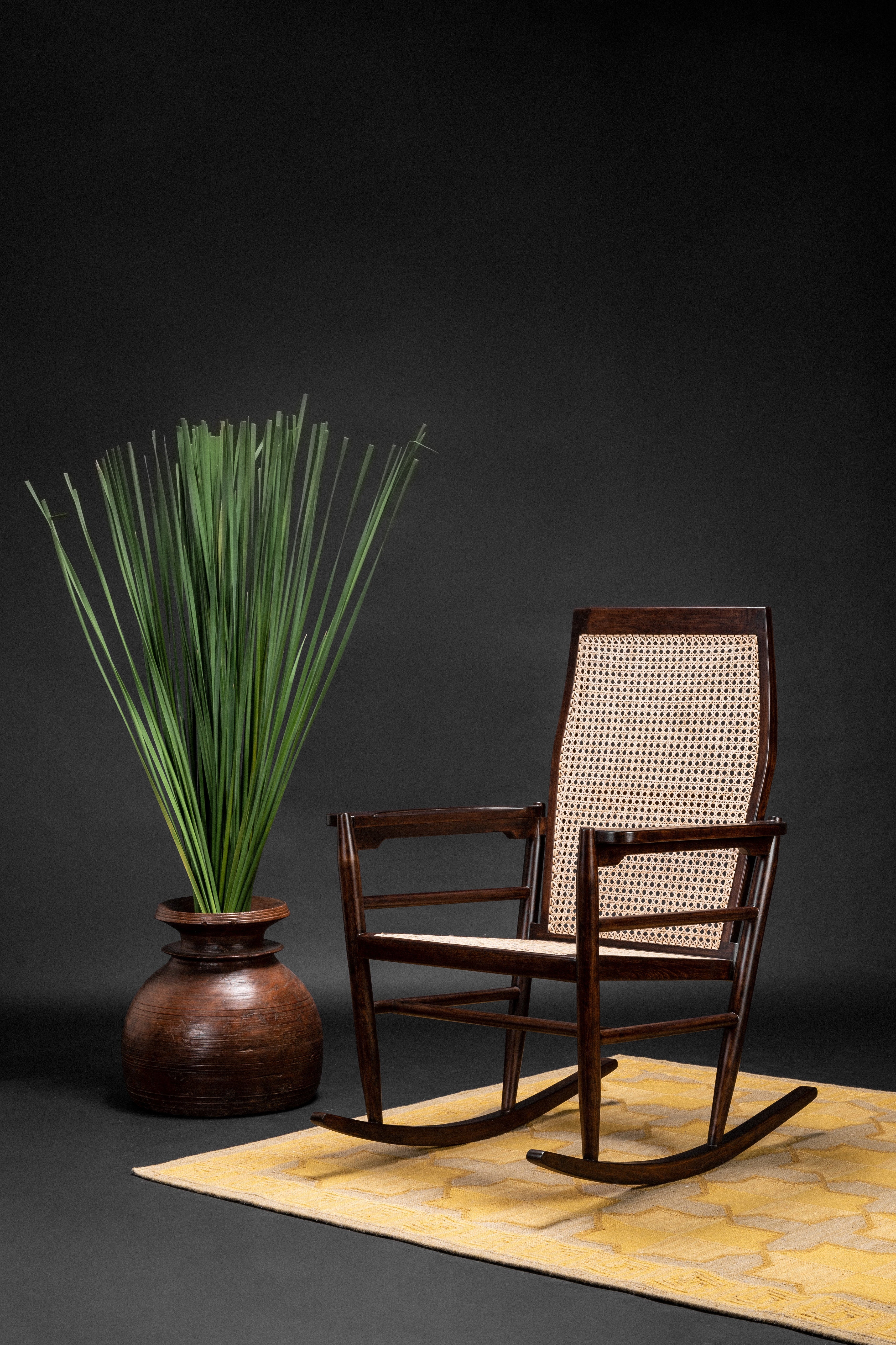 Wooden rocking chair with woven backrest next to a vase with greenery on a dark background