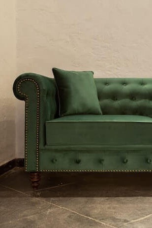 Green velvet sofa with tufted backrest and nailhead trim against a beige wall.