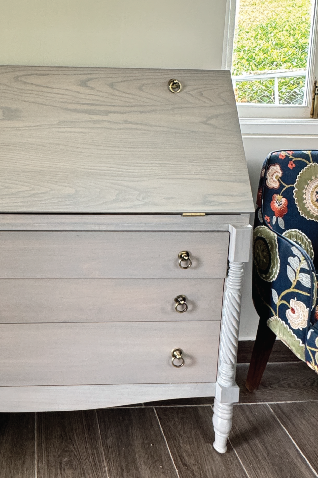 Wooden desk with drawers in a room with a window and floral-patterned chair.