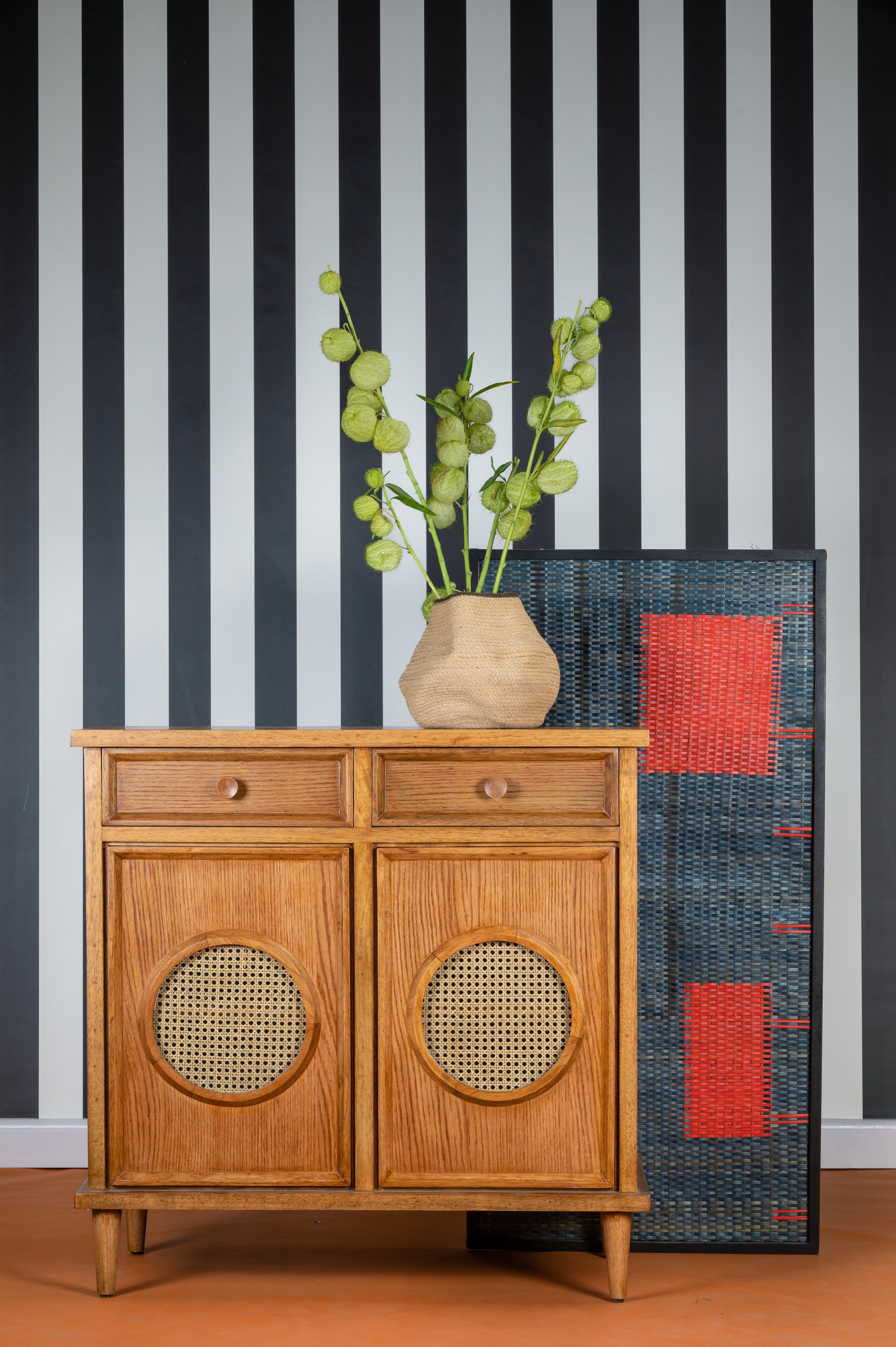 Wooden cabinet with a vase of greenery against a black and white striped wall.