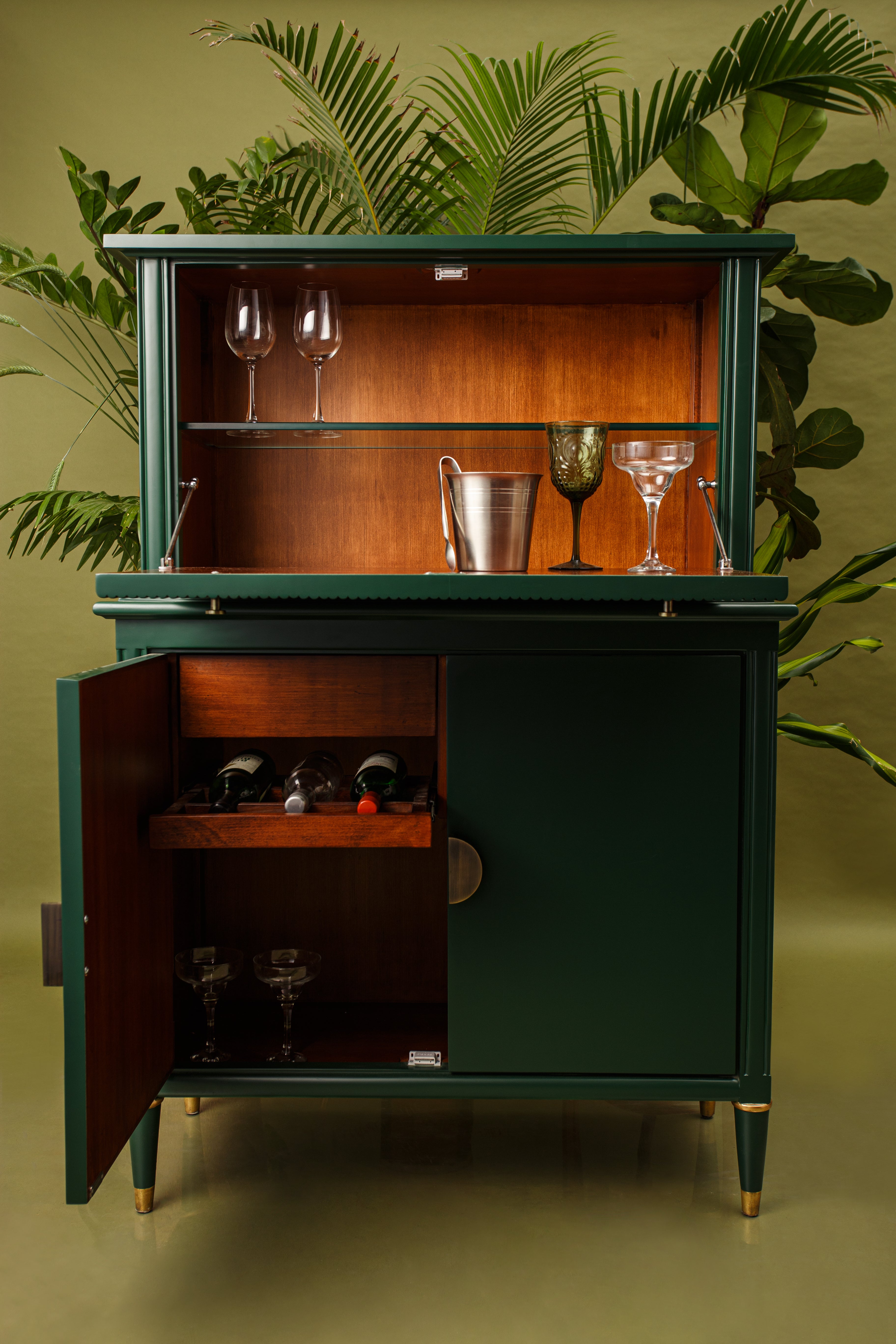 Green bar cabinet with open doors displaying wine glasses and bottles against a green background with plants.