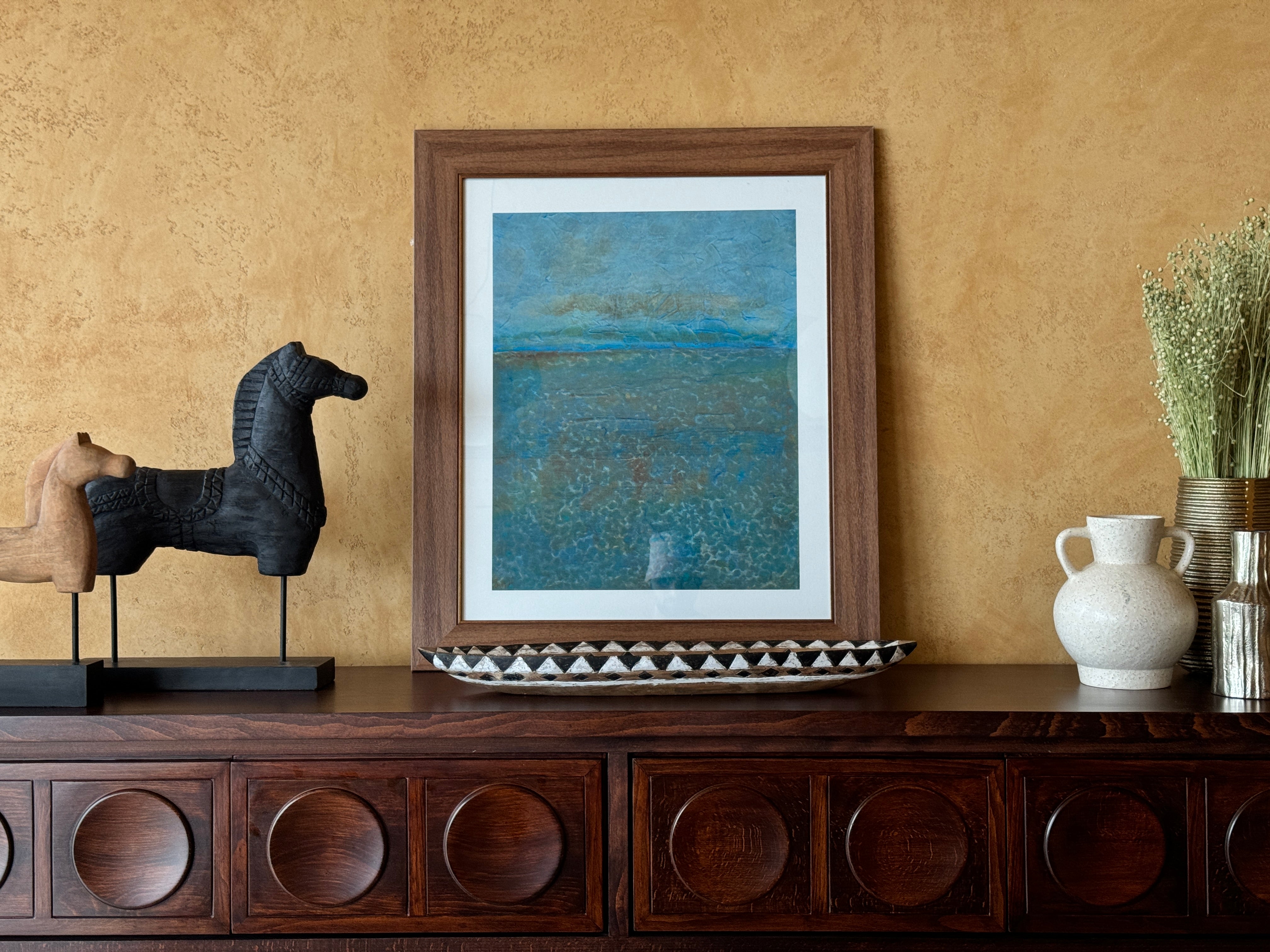 Framed artwork on a wooden console table with decorative items against a beige wall.