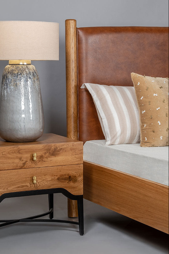Wooden nightstand with lamp next to a bed with pillows on a gray background