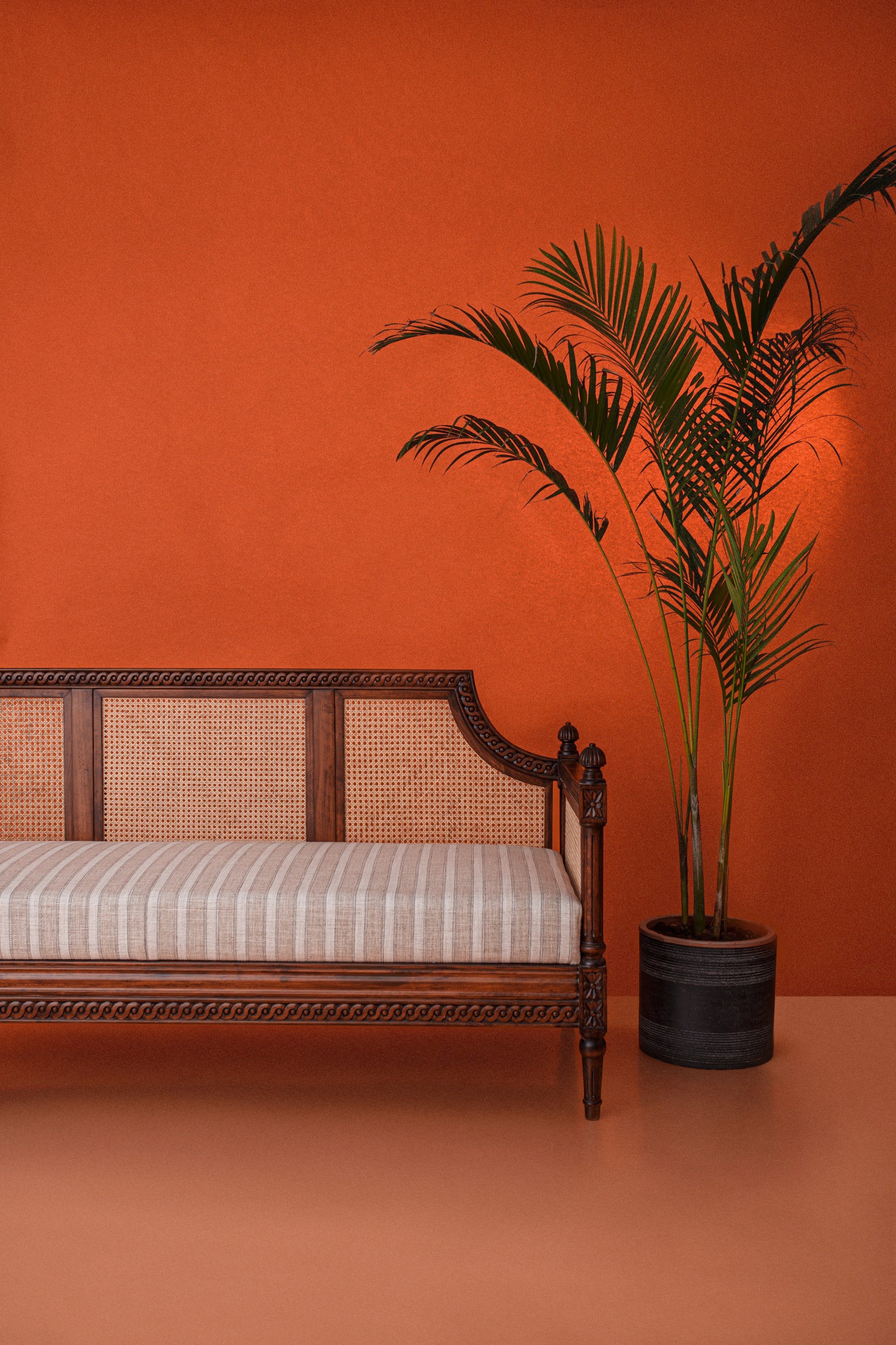 Wooden bench with striped cushion against an orange wall with a potted plant.