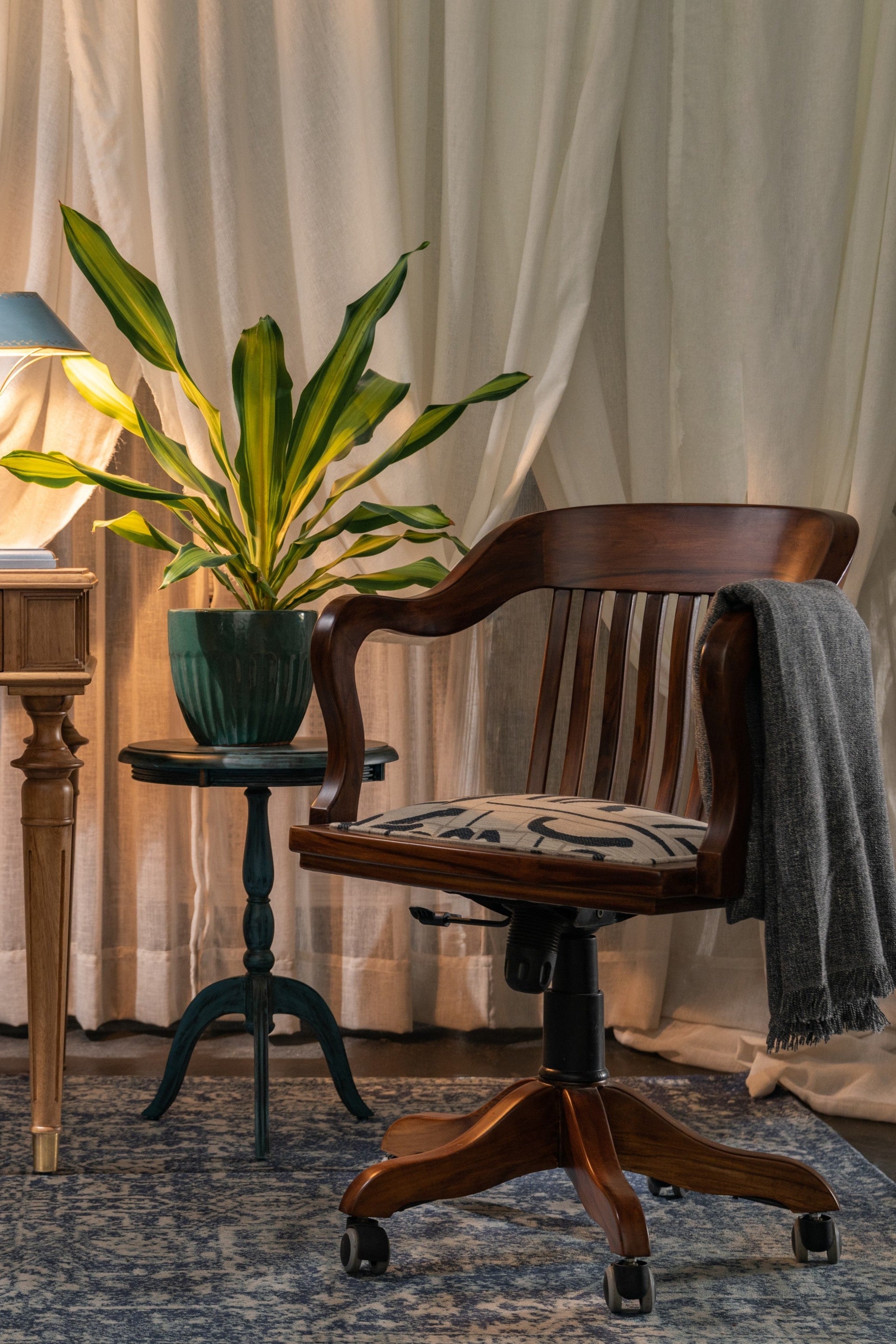 Vintage desk with lamp, plant, and chair in a room with white curtains and a rug.