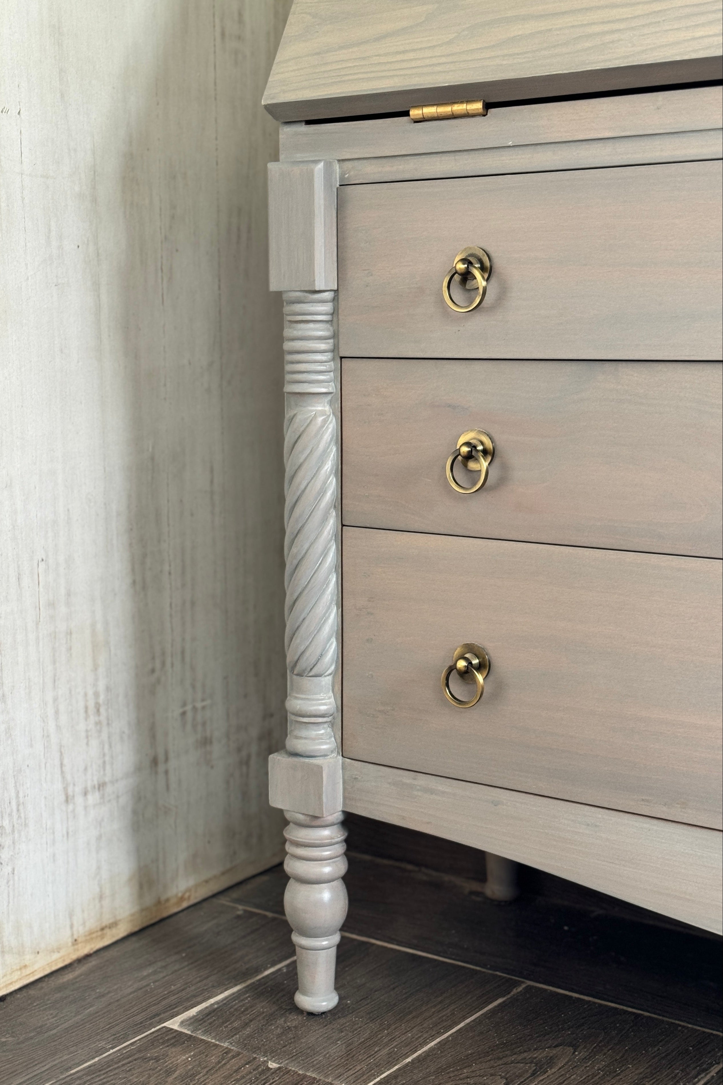 Wooden dresser with decorative leg and brass handles against a wooden wall.