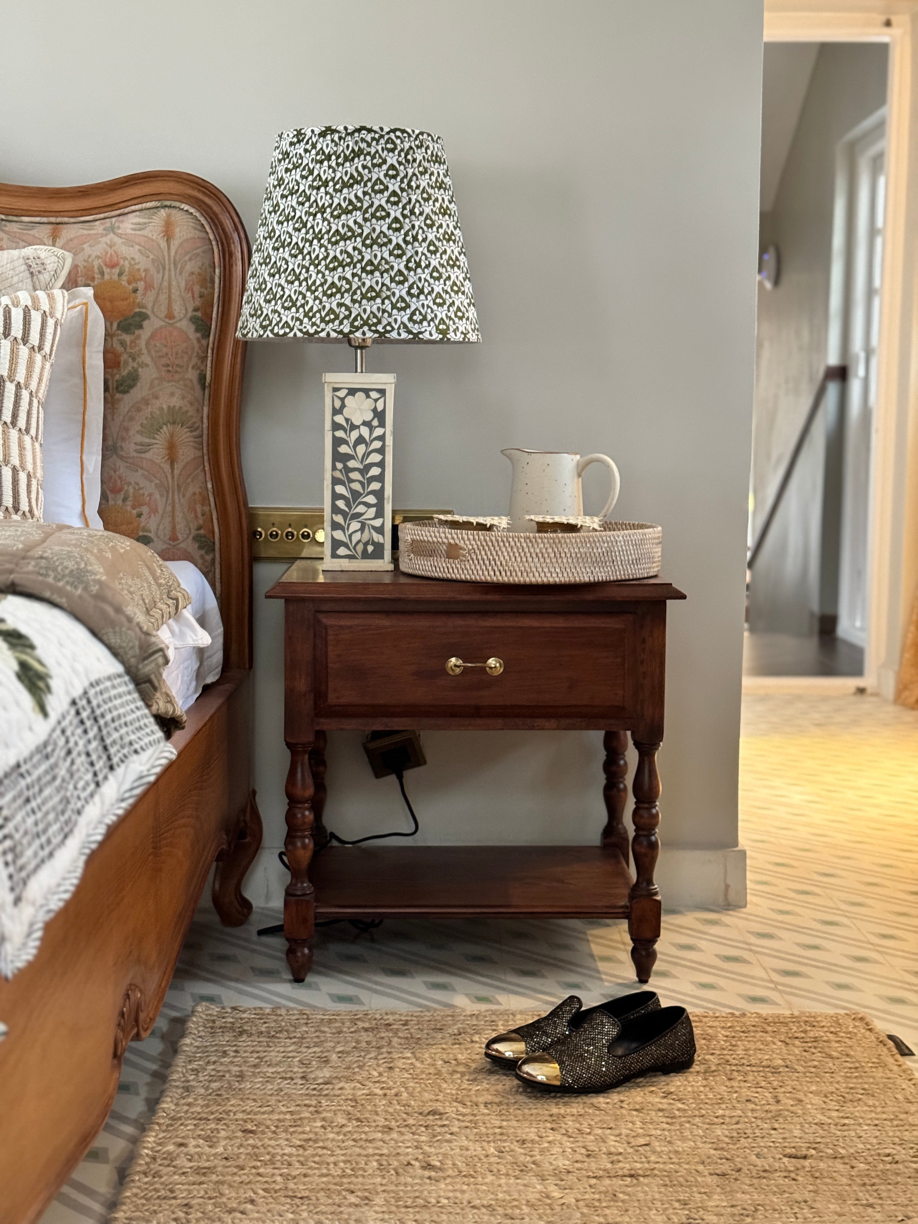 Wooden nightstand with lamp and decorative items next to a bed in a bedroom.