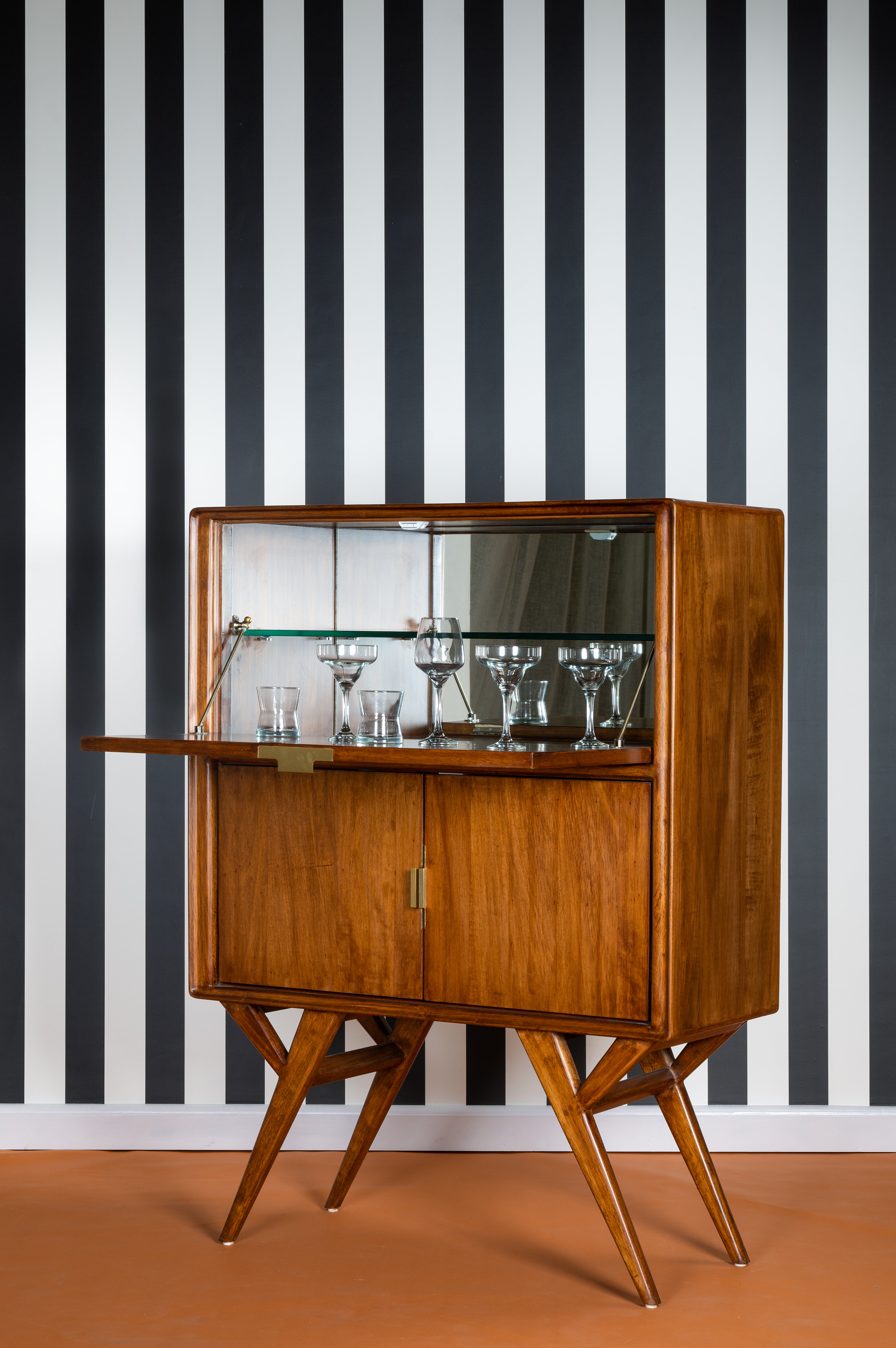 Wooden bar cabinet with glass shelves against a black and white striped wall.